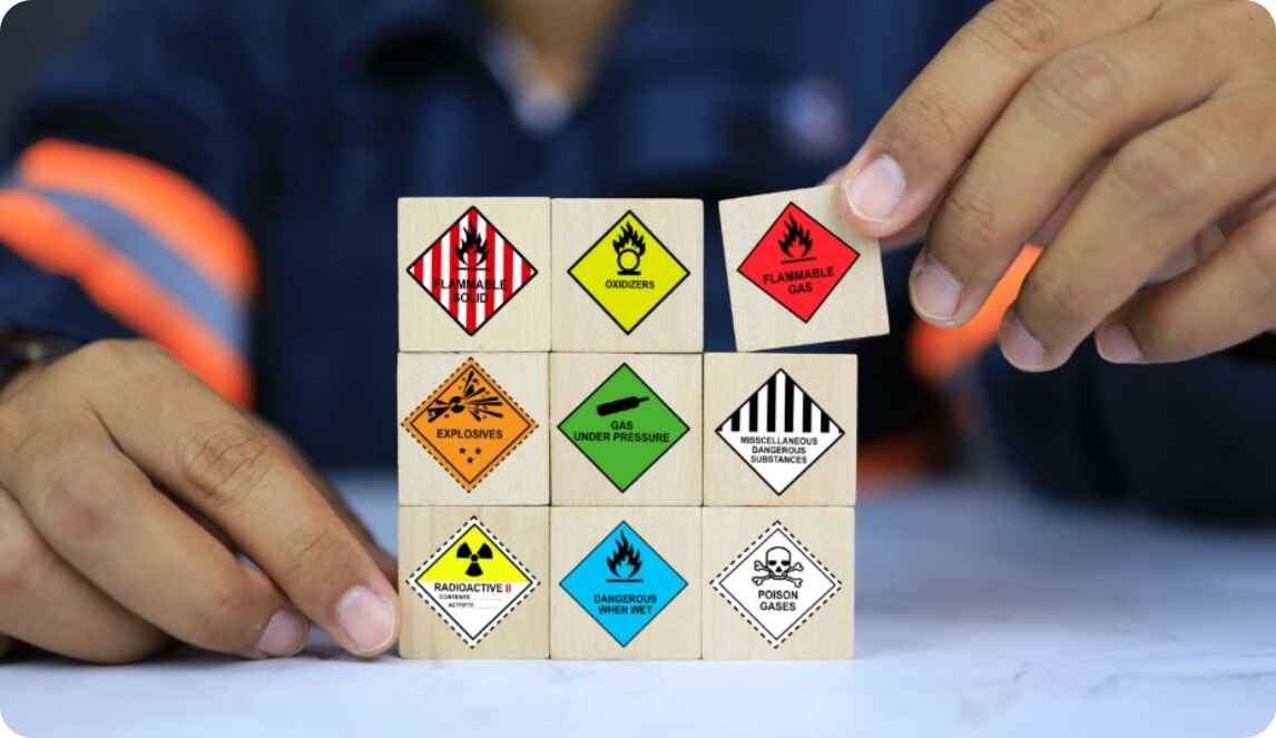 A person holds a wooden block displaying various safety signs related to dangerous goods shipping.